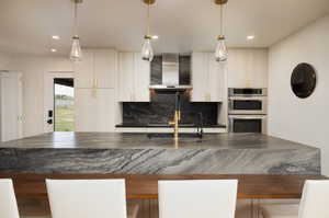 Kitchen featuring wall chimney exhaust hood, tasteful backsplash, pendant lighting, and double oven