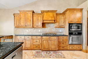 Kitchen with lofted ceiling, appliances with stainless steel finishes, premium range hood, dark stone counters, and tasteful backsplash