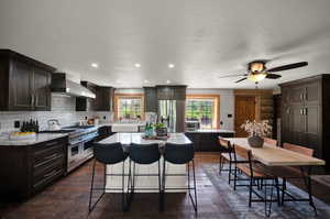 Kitchen with dark hardwood / real hardwood floors, ceiling fan, stainless steel appliances, a kitchen island, and wall chimney range hood