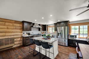 Kitchen featuring dark real hardwood flooring, a kitchen island, stainless steel appliances, wall chimney exhaust hood, and ceiling fan