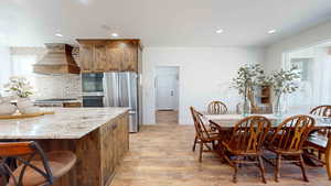 Kitchen with light stone countertops, light hardwood / wood-style flooring, custom exhaust hood, stainless steel appliances, and a breakfast bar