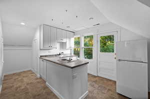 Kitchen with vaulted ceiling, white refrigerator, kitchen peninsula, tile patterned floors, and white cabinetry