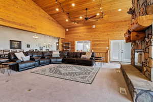 Carpeted living room with wooden walls, track lighting, wood ceiling, ceiling fan with notable chandelier, and high vaulted ceiling