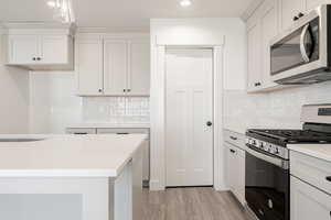 Kitchen with light wood-type flooring, backsplash, stainless steel appliances, white cabinetry, and hanging light fixtures