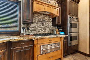 Kitchen featuring light tile patterned flooring, custom exhaust hood, decorative backsplash, light stone countertops, and appliances with stainless steel finishes