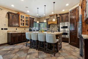 Kitchen featuring custom range hood, an island with sink, light stone countertops, stainless steel double oven, and a kitchen breakfast bar