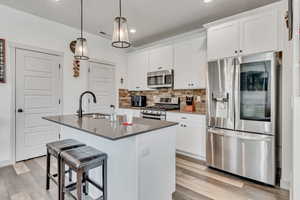 Kitchen featuring white cabinetry, an island with sink, stainless steel appliances, light hardwood / wood-style floors, and decorative backsplash