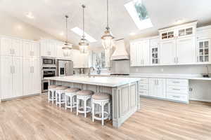 Kitchen with white cabinetry, light wood-type flooring, premium range hood, a kitchen island with sink, and stainless steel appliances