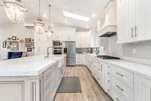 Kitchen featuring light hardwood / wood-style flooring, stainless steel appliances, hanging light fixtures, premium range hood, and a kitchen island with sink