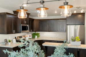 Kitchen featuring dark brown cabinetry, decorative light fixtures, stainless steel appliances, light stone countertops, and a textured ceiling