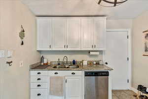 Kitchen with light tile patterned flooring, white cabinets, dark stone counters, stainless steel dishwasher, and sink