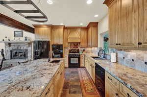 Kitchen with light stone countertops, black appliances, custom exhaust hood, a stone fireplace, and sink