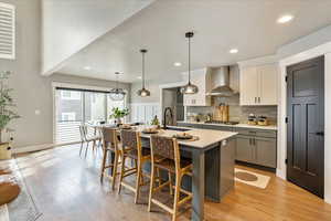 Kitchen featuring a kitchen island with sink, pendant lighting, wall chimney range hood, and light wood-type flooring