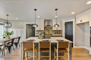 Kitchen with a center island with sink, wall chimney exhaust hood, light wood-type flooring, decorative light fixtures, and white cabinetry