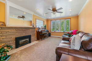 Living room with ornamental molding, carpet, a stone fireplace, and ceiling fan