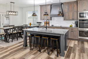 Kitchen featuring premium range hood, a kitchen island with sink, appliances with stainless steel finishes, hardwood / wood-style flooring, and decorative light fixtures