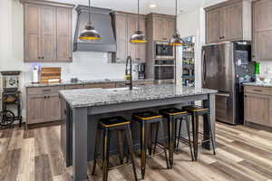 Kitchen with decorative light fixtures, an island with sink, custom range hood, light hardwood / wood-style floors, and appliances with stainless steel finishes