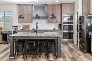 Kitchen with light wood-type flooring, light stone counters, stainless steel appliances, pendant lighting, and a center island with sink