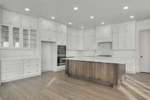 Kitchen with sink, light wood-type flooring, an island with sink, double oven, and white cabinetry