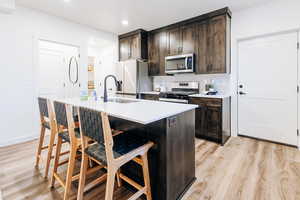 Basement Kitchen with stainless steel appliances, a center island with sink, sink, light hardwood / wood-style floors, and dark brown cabinetry