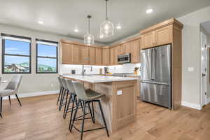 Kitchen featuring appliances with stainless steel finishes, light brown cabinets, hanging light fixtures, a kitchen island with sink, and light wood-type flooring