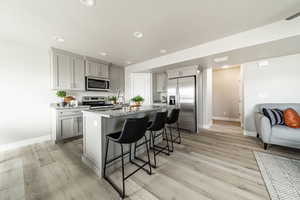 Kitchen with gray cabinets, light stone counters, appliances with stainless steel finishes, light hardwood / wood-style flooring, and a kitchen island with sink