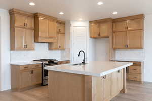 Kitchen with stainless steel gas stove, tasteful backsplash, light hardwood / wood-style floors, and an island with sink