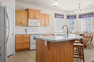 Kitchen with light tile patterned flooring, white appliances, hanging light fixtures, sink, and a center island with sink