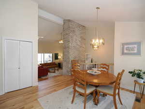 Dining area with ceiling fan with notable chandelier, vaulted ceiling, light hardwood / wood-style flooring, and brick wall