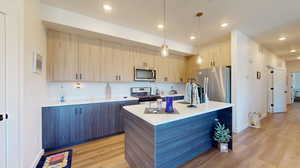 Kitchen with stainless steel appliances, pendant lighting, light brown cabinets, light hardwood / wood-style floors, and a center island