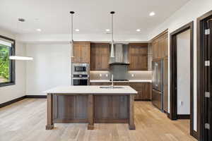 Kitchen with light hardwood / wood-style flooring, tasteful backsplash, hanging light fixtures, appliances with stainless steel finishes, and wall chimney exhaust hood