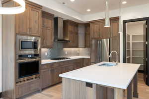 Kitchen with light wood-type flooring, a center island with sink, wall chimney range hood, and stainless steel appliances