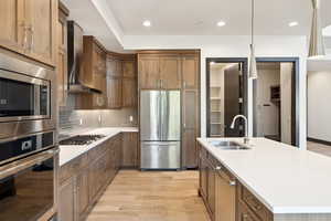 Kitchen featuring light hardwood / wood-style flooring, wall chimney range hood, an island with sink, appliances with stainless steel finishes, and sink