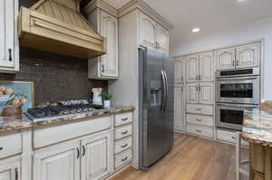 Kitchen with light wood-type flooring, light stone countertops, custom range hood, appliances with stainless steel finishes, and backsplash