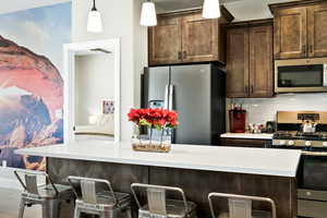 Kitchen featuring dark brown cabinets, stainless steel appliances, hanging light fixtures, a breakfast bar area, and backsplash