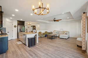 Interior space with light hardwood / wood-style flooring, ceiling fan with notable chandelier, a center island with sink, and a raised ceiling