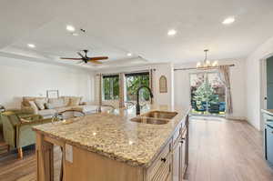 Kitchen with sink, a kitchen island with sink, light wood-type flooring, ceiling fan with notable chandelier, and a tray ceiling