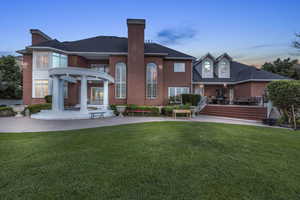 Back house at dusk featuring a wooden deck, a patio, a lawn, and a pergola