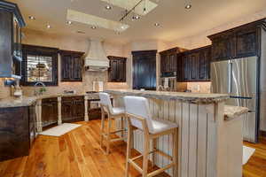 Kitchen featuring wood-style flooring, custom range hood, stainless steel appliances, and dark brown cabinets