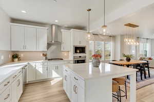 Kitchen with light hardwood / wood-style flooring, white cabinets, appliances with stainless steel finishes, wall chimney exhaust hood, and decorative backsplash