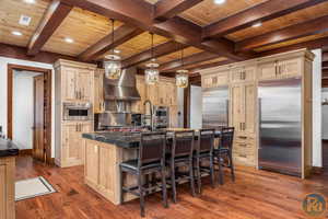 Kitchen featuring appliances with stainless steel finishes, a center island with sink, wall chimney range hood, light brown cabinets, and beam ceiling