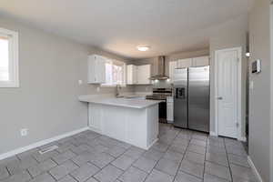 Kitchen featuring appliances with stainless steel finishes, kitchen peninsula, light tile patterned floors, white cabinetry, and wall chimney exhaust hood