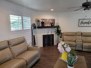 Living room featuring ceiling fan and dark wood-type flooring, and cozy fireplace.