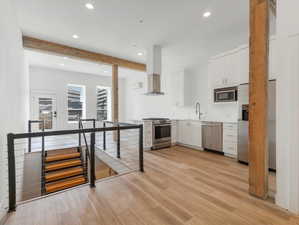 Kitchen featuring stainless steel appliances, white cabinets, wall chimney exhaust hood, beam ceiling, and light hardwood / wood-style floors