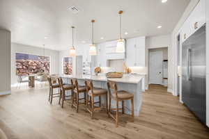 Kitchen featuring appliances with stainless steel finishes, light wood-type flooring, a large island with sink, white cabinets, and hanging light fixtures