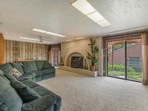 Living room featuring wooden walls, carpet, a textured ceiling, a brick fireplace, and ceiling fan