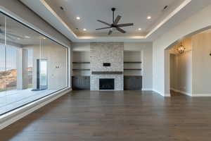 Unfurnished living room featuring a stone fireplace, a raised ceiling, built in features, and ceiling fan