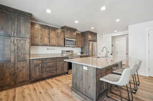 Kitchen with light wood-type flooring, a kitchen breakfast bar, stainless steel appliances, a kitchen island with sink, and light stone countertops