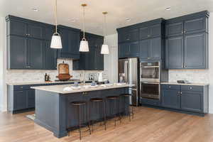 Kitchen with decorative backsplash, light hardwood / wood-style flooring, pendant lighting, an island with sink, and stainless steel appliances