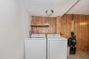 Laundry area with washing machine and clothes dryer, a textured ceiling, wooden walls, and light hardwood / wood-style floors